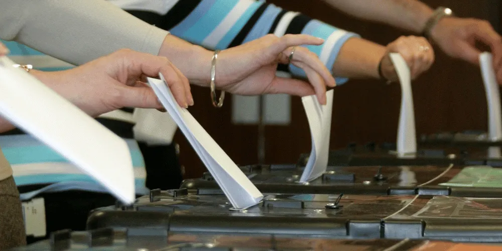 Four people's hands dropping ballot papers into ballot boxes at an election.