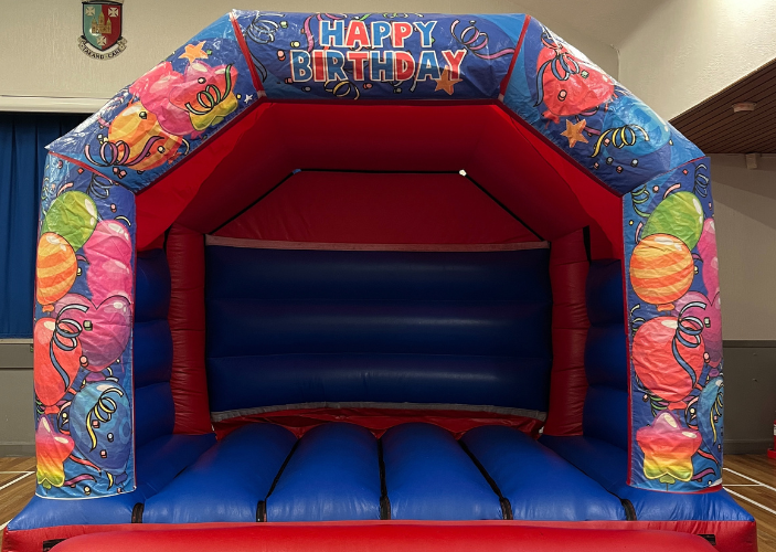 Blue and red bouncy castle with decorative images of balloons and Happy Birthday signage, in community hall