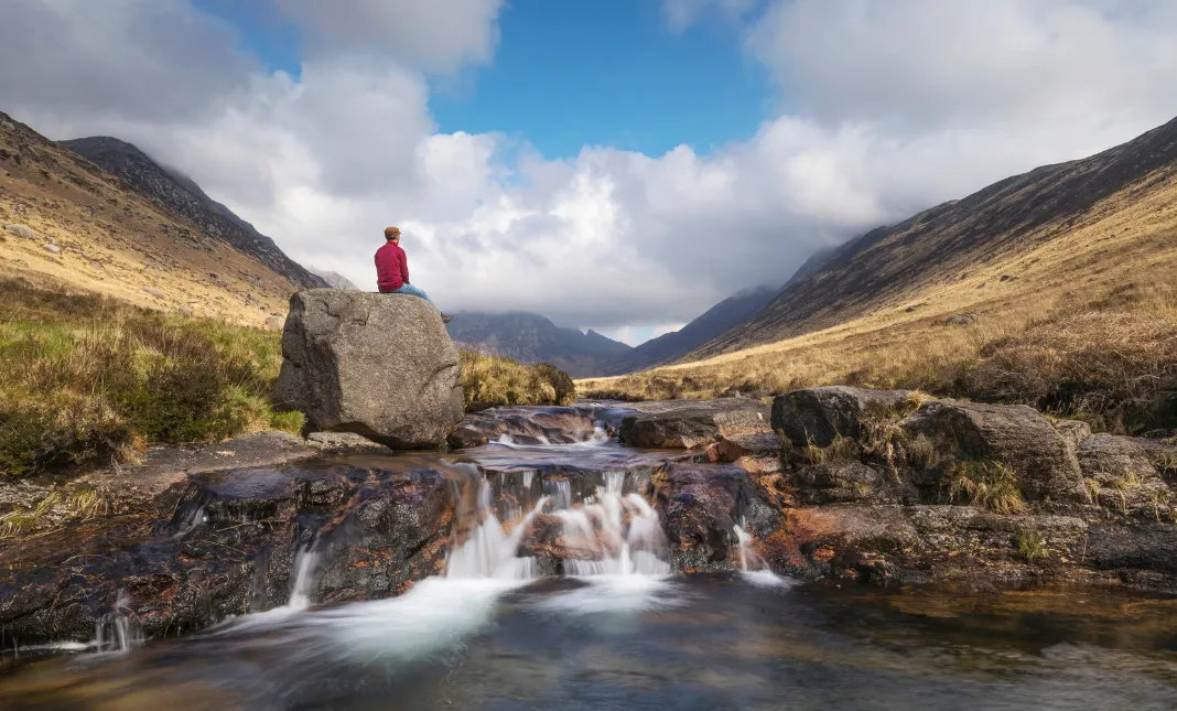 Person wearing a red top sits on top of a large boulder by a stream in a glen