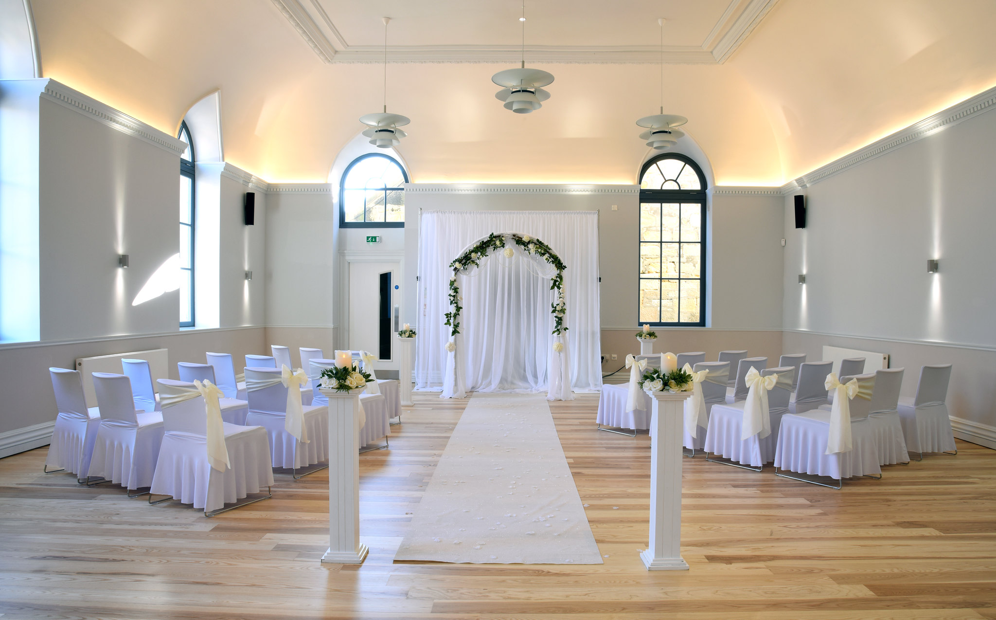 Room with arched windows set up for wedding with draped chairs, arch and flowers